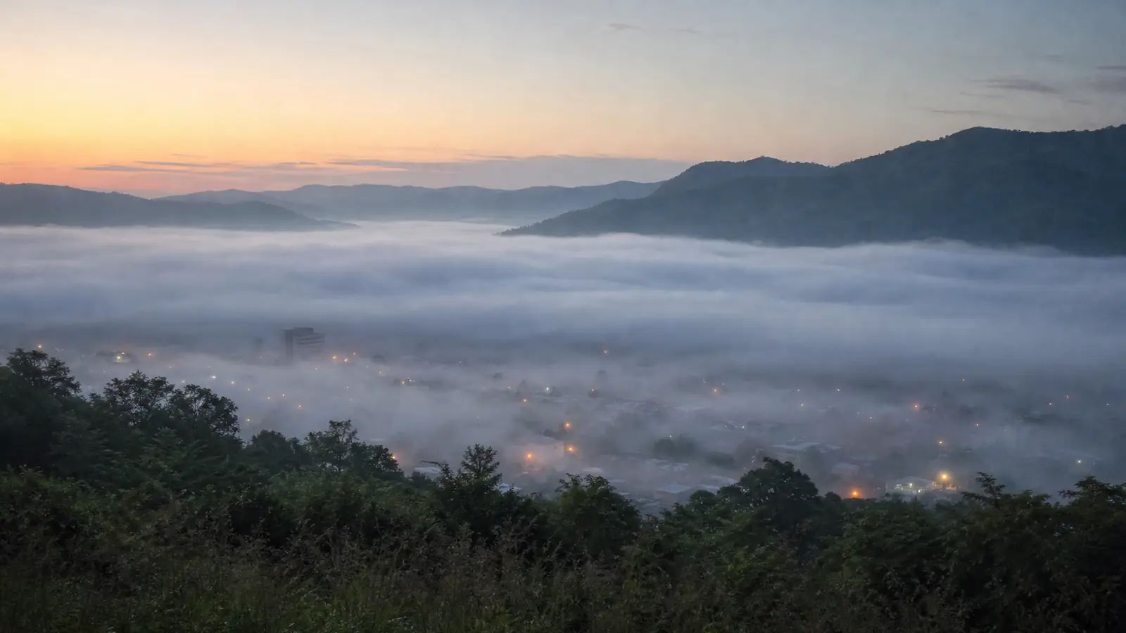 A small city in a foggy valley at dawn, with low cloud lying between muted grey-green hills and soft rooftop lights.