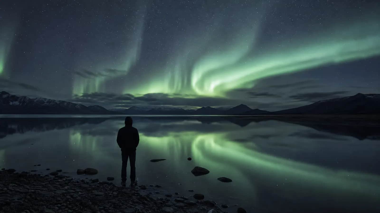 Person in a hood standing on a lake shore, green aurora reflecting on water, mountains in background