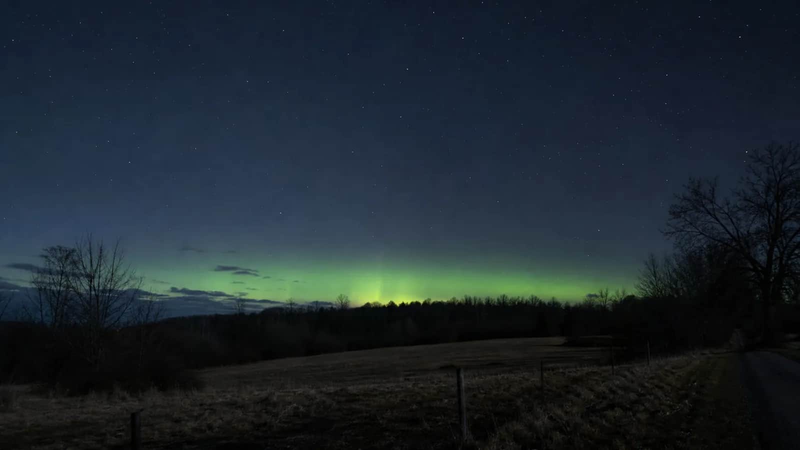 Faint green arc of aurora above a treeline, field and fence post under a starry sky