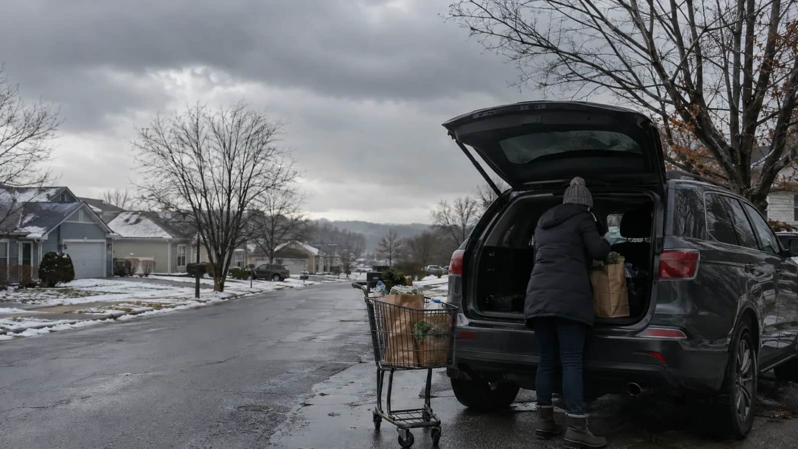 A grey, overcast sky sits over a suburban street where people load grocery bags into a car before an expected winter storm.